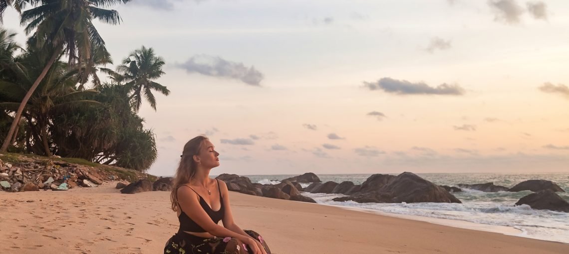 Alone young woman sitting on sandy beach at palm trees and sea background, relaxing, thinking looking away. Pretty lady wear black dress on tropical ocean. Travel vacation concept. Copy text space